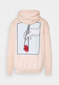 Sudadera beige con gráfico de una mano sosteniendo un corazón anatómico rojo con palillos, una sola gota cayendo y el texto "VAS A ESTAR BIEN."