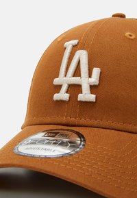 Gorra marrón con el logo bordado de "LA" en blanco. Presenta una tela texturizada, correa ajustable y una etiqueta circular de la marca en la visera.