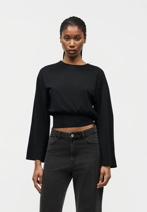 Giovane donna con capelli intrecciati che indossa un top nero a manica lunga e pantaloni a vita alta grigio scuro, in piedi contro uno sfondo chiaro.