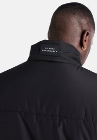 Svart isolerad jacka gjord av slät tyg, med hög krage och en märkestagg på baksidan. Enkel design utan ytterligare detaljer.