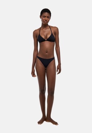 Femme debout pieds nus portant un bikini noir deux pièces sur un fond blanc uni, face à l'avant avec une expression neutre.