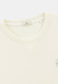 Sweatshirt couleur crème en tissu doux, avec un col rond et une texture côtelée. Présente une petite étiquette de logo près de l'ourlet et des accents de couture.