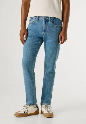 Hellblaue Jeans mit geradem Schnitt, fünf Taschen und kontrastierender Naht, getragen mit weißen und beigen Turnschuhen.