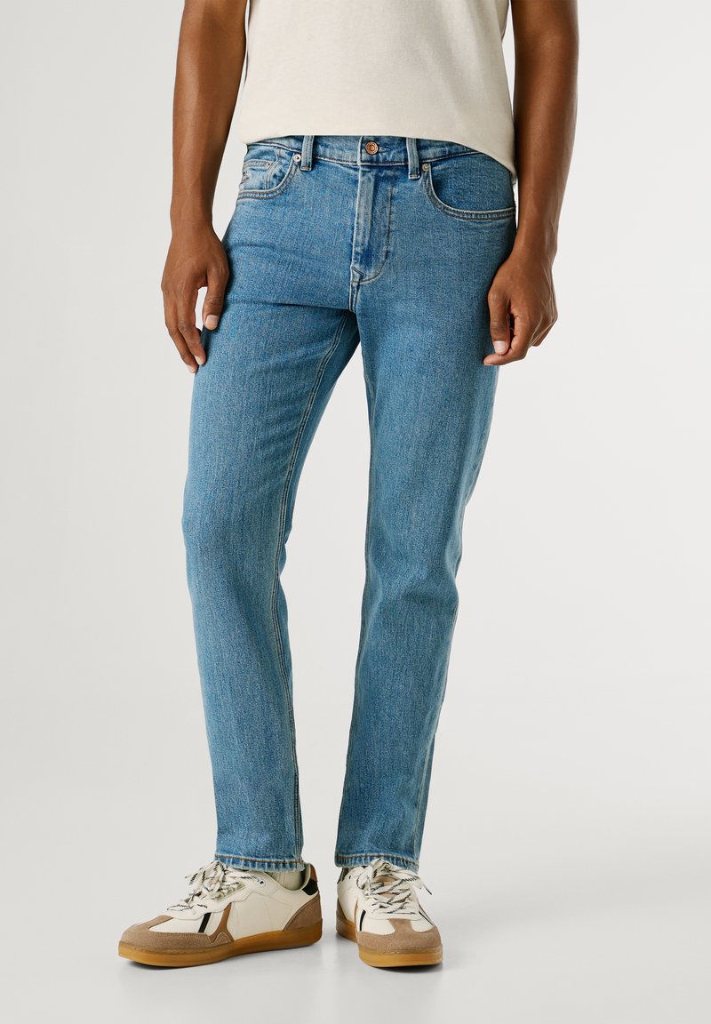 Jasnoniebieskie dżinsy o prostym kroju, z pięcioma kieszeniami i kontrastowym szwem, noszone z białymi i beżowymi trampkami.