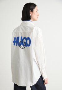 Hvit langermet skjorte med blå spraymalt "HUGO"-tekst og et grafisk smilende ansikt på baksiden, med klassisk krage og knappe mansjetter.