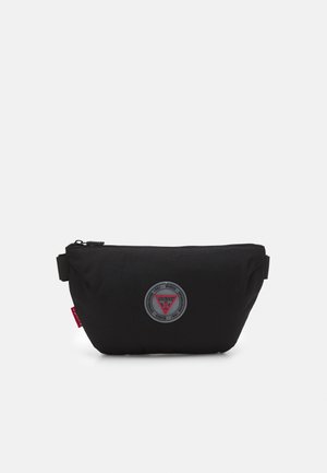 Zwarte stoffen heuptas met rits, cirkelvormige Guess-logo patch in het midden en een rood label aan de linkerkant tegen een witte achtergrond.