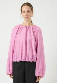 Lyserød bluse med puffærmer lavet af skinnende stof, med en rynket halsudskæring og elastisk kant, kombineret med sorte bukser.