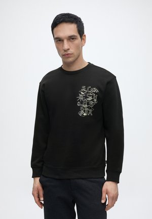 Mann trägt schwarzen Sweatshirt mit abstraktem weißen Grafiktext auf der linken Brust, steht vor schlichtem hellem Hintergrund.