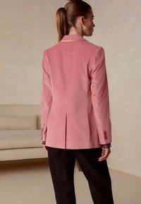 Pink corduroy blazer with a structured design, single vent at the back, and button accents on the sleeves. Worn over dark pants.