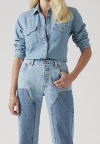 Vaaleansininen farkkupaita rintataskuilla, yhdistetty korkeavyötäröisiin vaaleansinisiin farkkuihin, joissa on kontrastisia applikaatiodetaljeja ja näkyvät saumat.