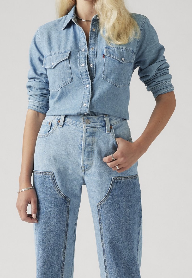 Vaaleansininen farkkupaita rintataskuilla, yhdistetty korkeavyötäröisiin vaaleansinisiin farkkuihin, joissa on kontrastisia applikaatiodetaljeja ja näkyvät saumat.