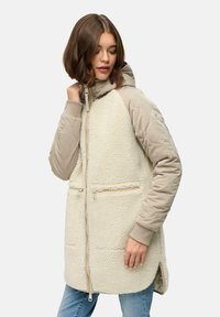Beige hættejakke med quiltet overdel og fluffy tekstureret nederste del, der har lynlås hele vejen ned foran og to forlommer.