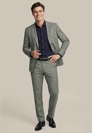 Junger Mann lächelt, trägt einen hellgrauen Anzug mit schwarzem Gürtel, dunkelblaues Hemd und schwarze Anzugsschuhe, steht vor einem schlichten Hintergrund.