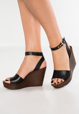 Czarne skórzane sandały na koturnie z odkrytym palcem, regulowanym paskiem na kostkę oraz brązową platformą z rowkowanym spodem, zapewniającą dodatkową wysokość i stabilność.