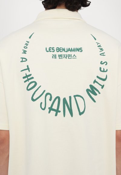 Polo shirt couleur crème avec un texte vert au dos : "LES BENJAMINS" et "FROM A THOUSAND MILES AWAY" disposés en cercle.