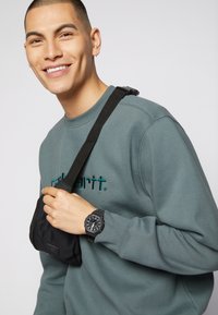 Black bag worn crossbody, gray sweatshirt with teal embroidery, black wristwatch with large face and textured strap, light background.
