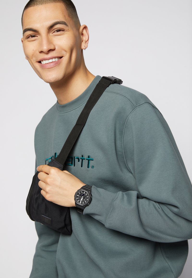 Black bag worn crossbody, gray sweatshirt with teal embroidery, black wristwatch with large face and textured strap, light background.