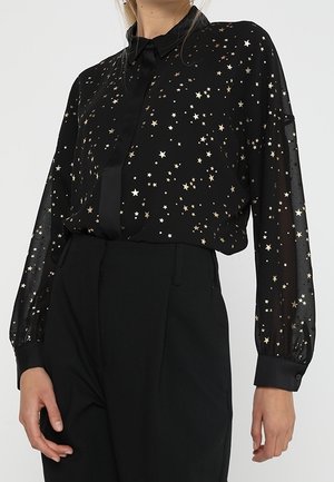 Blouse noire transparente avec motif d'étoiles dorées, poignets boutonnés et col noir, associée à un pantalon noir taille haute.