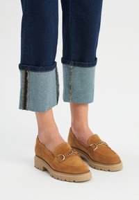 Suede loafers w kolorze tan z akcentem w postaci łańcuszka, w zestawieniu z niebieskimi dżinsami z kontrastowymi mankietami i postrzępionymi brzegami, podkreślonymi grubą podeszwą.