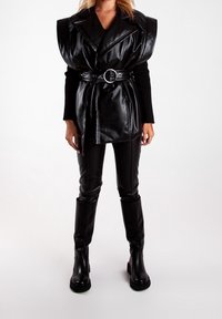 Femme portant un gilet en cuir sans manches noir avec des épaules larges, une taille ceinturée, un top à manches longues noir, un pantalon en cuir et des bottes noires sur fond blanc.