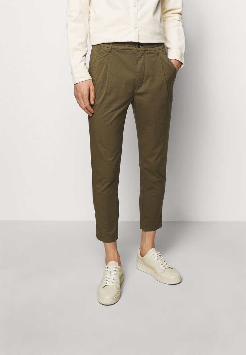 Homme portant un pantalon olive coupé, une chemise blanche boutonnée et des baskets beige clair, debout devant un fond uni.