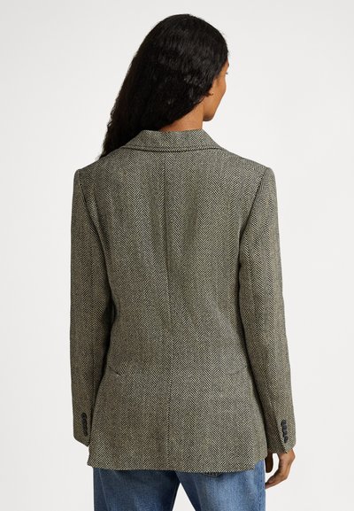 Kobieta o długich ciemnych włosach, ubrana w szarą marynarkę w jodełkę i niebieskie dżinsy, widziana od tyłu na białym tle.