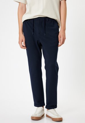 Un pantalon de survêtement bleu marine avec une taille élastique, un cordon de serrage et des poches latérales. Le tissu est doux, avec une coupe décontractée, associé à des baskets blanches.