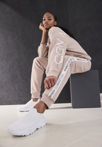 Beige sweatshirt en joggers hebben witte tekst langs de zijkanten. Het model draagt witte instap schoenen, zittend op een grijze kubus tegen een donkere achtergrond.