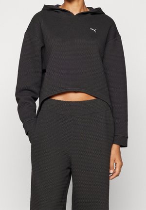 Personne portant un sweat à capuche court noir texturé et un pantalon taille haute assorti avec un petit logo Puma blanc sur la poitrine.