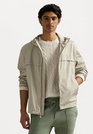 Licht beige lichtgewicht jack met een capuchon, twee borstzakken en elastische manchetten, gelaagd over een witte kabeltrui en groene pants.