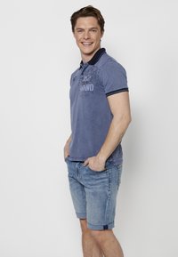 Moška modra polo majica z teksturirano površino, ki ima temen ovratnik, kratke rokave in majhne logotipne detajle. V kombinaciji z svetlim jeans kratkimi hlačami.