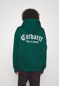 Sudadera con capucha verde hecha de tela suave que presenta el logo "Carhartt" y el texto "Work in Progress" en gran impresión blanca en la parte trasera.