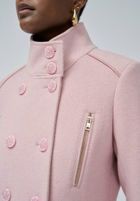 Abrigo de espiga rosa con un cuello alto, seis botones grandes y un detalle de bolsillo con cremallera en el lateral. Textura suave con un diseño estructurado.