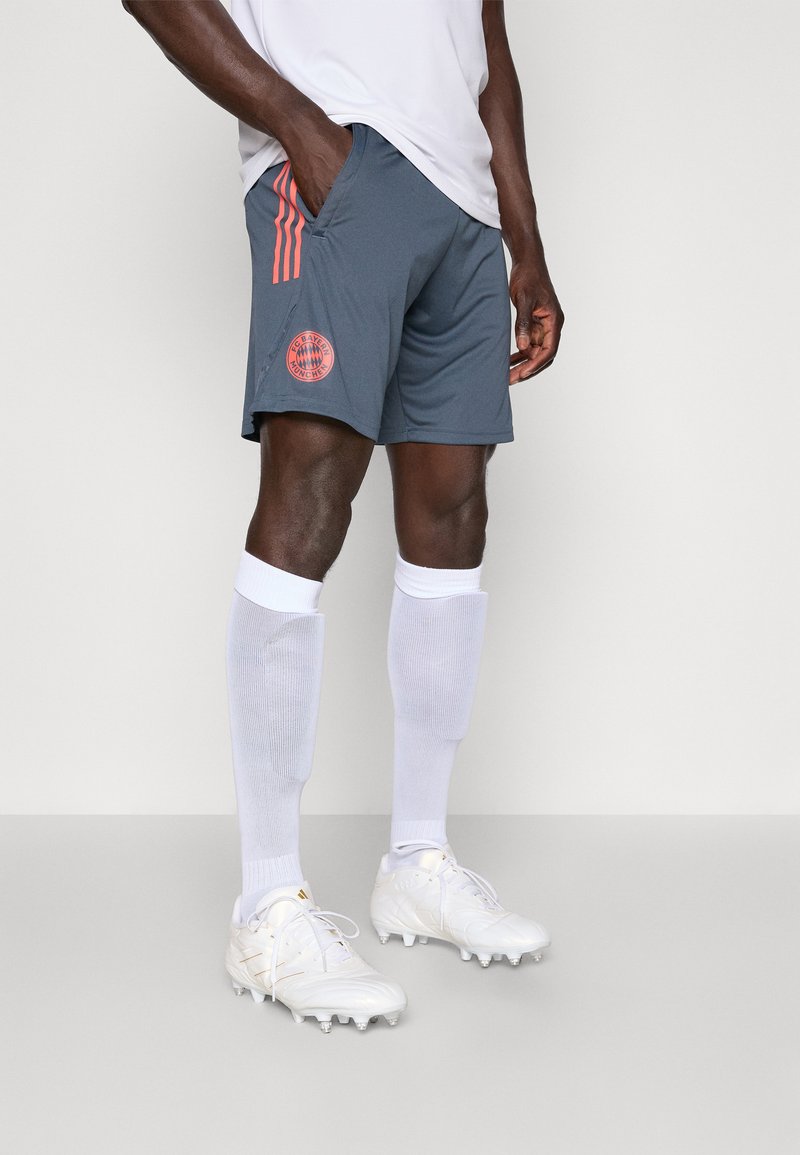 Shorts de sport gris avec trois rayures orange et un logo rond. Associés à des chaussettes blanches montant au genou et des chaussures blanches avec des accents dorés.