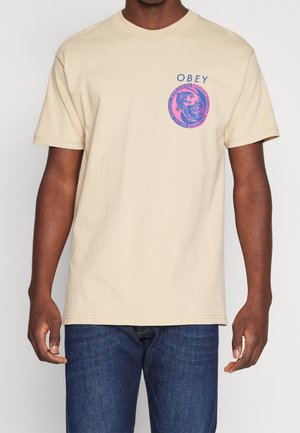 Beżowa bawełniana koszulka z krótkim rękawem z okrągłym logo w różowym i niebieskim napisie "OBEY". Standardowy krój.