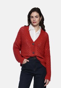 Roter Strickcardigan mit lockerem Schnitt, V-Ausschnitt und Knopfleiste. Verfügt über ein offenes Muster und gerippte Bündchen. Kombiniert mit dunkelblauen Jeans.