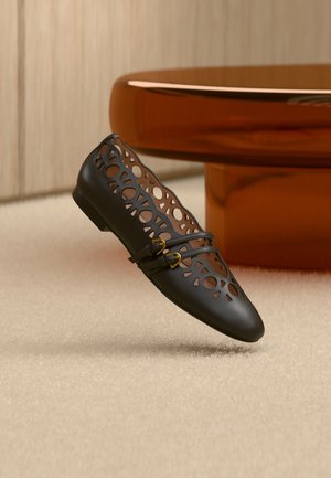 Μαύρο δερμάτινο μποτάκι με γεωμετρικές εγκοπές και δύο μικρά χρυσά λουράκια με αγκράφες, τοποθετημένο πάνω σε μπεζ χαλί και κοντά σε ένα καφέ στρογγυλό τραπέζι.