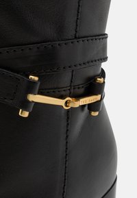 Botte en cuir noir avec une texture lisse, dotée d'une boucle dorée, de couture élaborée et d'une accent de sangle étroite.