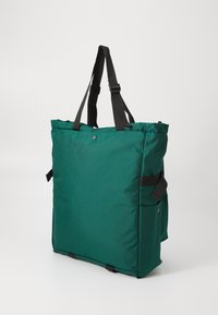 Grande bolsa tote verde feita de um tecido durável, com alças pretas ajustáveis, um design minimalista e bolsos laterais para armazenamento adicional.