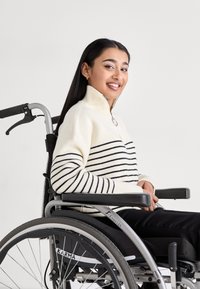 Maglione crema con strisce blu navy, colletto alto e texture a costine indossato da una persona in una sedia a rotelle argento con dettagli neri e un logo.