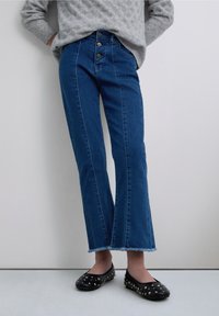 Blauwe denim jeans met een hoge taille en een rechte pijp, een rafelige zoom en contrasterende stiksels, gecombineerd met zwarte platte schoenen met studs.