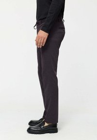 Man standing in dark purple trousers, black long-sleeve shirt, and black loafers against a neutral background.