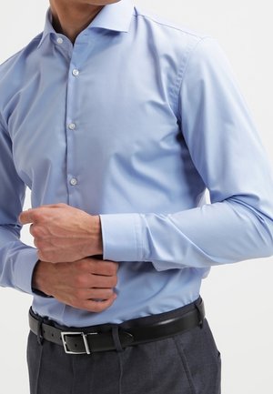 Man in light blue buttoned dress shirt adjusting his cuff, wearing dark trousers and a black belt, against a plain background.