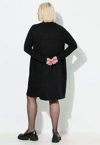 Sort langærmet kjole med en tætsiddende overdel og samlet talje. Har en glat tekstur og falder over knæet, parret med chunky sorte sko.