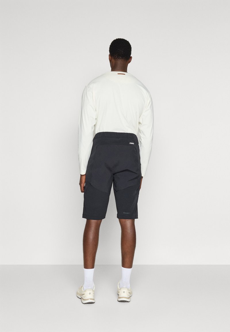 Mann trägt ein weißes, langärmliges Hemd, schwarze Shorts, weiße Socken und beigefarbene Turnschuhe und steht mit dem Gesicht zu einer schlichten hellgrauen Wand.