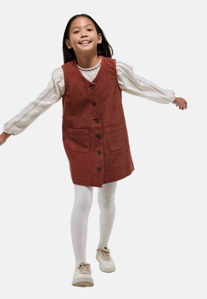 Robe en velours côtelé couleur rouille avec des boutons sur le devant, deux poches et un décolleté en V. Associée à un haut à manches longues blanc et des collants blancs.