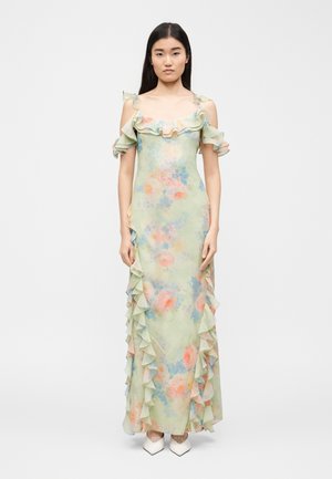 Vrouw staat, draagt een pastelgekleurde bloemenprint maxi-jurk met cold-shoulder en gerimpelde randen, gecombineerd met witte pumps met scherpe neus, tegen een effen achtergrond.