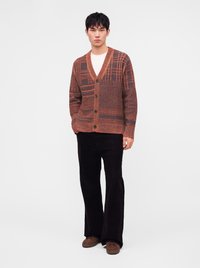 Homme portant un cardigan marron à motifs boutonné, une chemise blanche, un pantalon noir large et des chaussures marron, debout devant un fond uni.