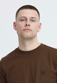 Smeđa pamučna majica s okruglim izrezom i kratkim rukavima; sadrži mali crni izvezeni logo na lijevoj strani prsnog koša.