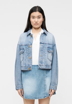 Frau trägt eine hellblaue, kurz geschnittene Jeansjacke mit silbernen Knöpfen über einem weißen Hemd, kombiniert mit einem passenden Jeansrock vor einfachem Hintergrund.